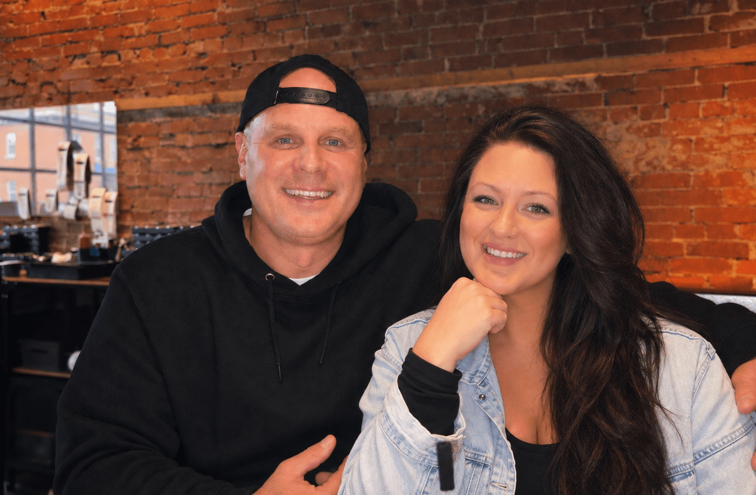 Smiling man in black hoodie and woman in denim jacket pose before red brick wall.